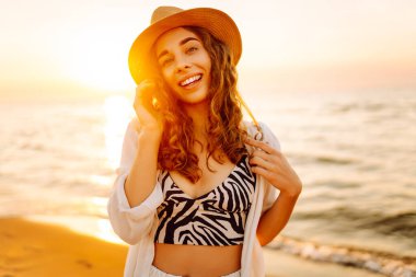 Güneş batarken sahilde eğlenen şapkalı genç bir kadının portresi. Güzel bayan turist özgürlüğü hissediyor ve dışarıda vakit geçirmeyi seviyor. Yaşam tarzı, eğlence..