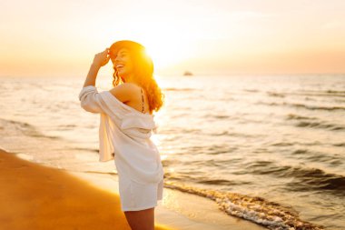 Güneş batarken sahilde eğlenen şapkalı genç bir kadının portresi. Güzel bayan turist özgürlüğü hissediyor ve dışarıda vakit geçirmeyi seviyor. Yaşam tarzı, eğlence..