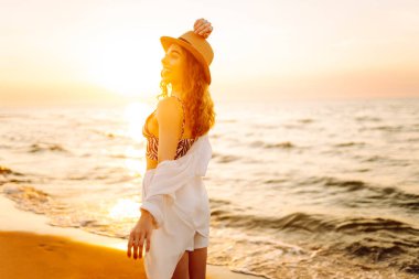 Güneş batarken sahilde eğlenen şapkalı genç bir kadının portresi. Güzel bayan turist özgürlüğü hissediyor ve dışarıda vakit geçirmeyi seviyor. Yaşam tarzı, eğlence..