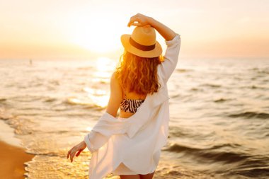 Güneş batarken sahilde eğlenen şapkalı genç bir kadının portresi. Güzel bayan turist özgürlüğü hissediyor ve dışarıda vakit geçirmeyi seviyor. Yaşam tarzı, eğlence..