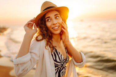 Güneş batarken sahilde eğlenen şapkalı genç bir kadının portresi. Güzel bayan turist özgürlüğü hissediyor ve dışarıda vakit geçirmeyi seviyor. Yaşam tarzı, eğlence..