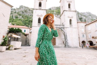 Parlak elbiseli ve şapkalı güzel bir turist eğleniyor ve şehir manzarasının tadını çıkarıyor. Genç bir kadın şehirde geziyor ve eğleniyor. Tatil, tatil, seyahat konsepti.