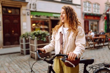 Gündelik giysiler içinde güzel bir kadın güneşli şehir sokaklarında bisiklet sürüyor. Yürüyüş, tatil, seyahat kavramı. Etkin yaşam biçimi.