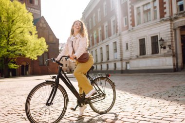 Gündelik giysiler içinde güzel bir kadın güneşli şehir sokaklarında bisiklet sürüyor. Yürüyüş, tatil, seyahat kavramı. Etkin yaşam biçimi.