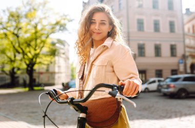 Gündelik giysiler içinde güzel bir kadın güneşli şehir sokaklarında bisiklet sürüyor. Yürüyüş, tatil, seyahat kavramı. Etkin yaşam biçimi.