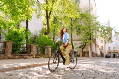 Gündelik giysiler içinde güzel bir kadın güneşli şehir sokaklarında bisiklet sürüyor. Yürüyüş, tatil, seyahat kavramı. Etkin yaşam biçimi.