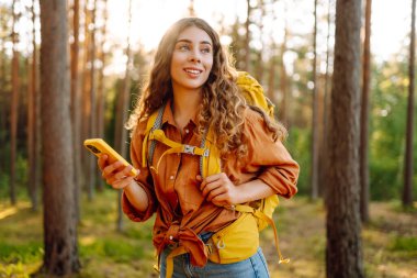 Sırt çantalı ve elinde telefon olan sevimli bir bayan gezgin güneşli bir ormanda yürüyor. Güzel bayan turist blogları ve gün batımının tadını çıkarıyor. Etkin yaşam biçimi.