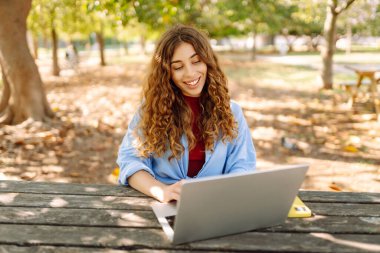 Parktaki bir masada dizüstü bilgisayarla çalışan genç bir kadın. Açık havada havanın tadını çıkaran mutlu kadın serbest yazar. Uzaktan çalışma konsepti. Yaşam biçimi.