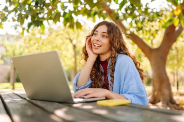 Parktaki bir masada dizüstü bilgisayarla çalışan genç bir kadın. Açık havada havanın tadını çıkaran mutlu kadın serbest yazar. Uzaktan çalışma konsepti. Yaşam biçimi.