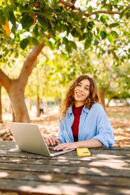 Parktaki bir masada dizüstü bilgisayarla çalışan genç bir kadın. Açık havada havanın tadını çıkaran mutlu kadın serbest yazar. Uzaktan çalışma konsepti. Yaşam biçimi.