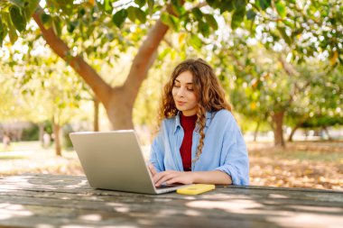 Parktaki bir masada dizüstü bilgisayarla çalışan genç bir kadın. Açık havada havanın tadını çıkaran mutlu kadın serbest yazar. Uzaktan çalışma konsepti. Yaşam biçimi.