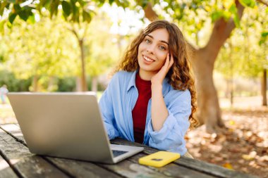 Parktaki bir masada dizüstü bilgisayarla çalışan genç bir kadın. Açık havada havanın tadını çıkaran mutlu kadın serbest yazar. Uzaktan çalışma konsepti. Yaşam biçimi.