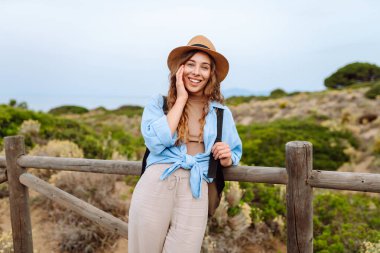 Sırt çantalı, şapkalı, ahşap patikada yürüyen genç bir kadın gezginin arka görüntüsü. Doğanın tadını çıkaran güzel bir kadın, deniz manzarası. Yürüyüş, yürüyüş konsepti.