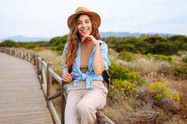 Sırt çantalı, şapkalı, ahşap patikada yürüyen genç bir kadın gezginin arka görüntüsü. Doğanın tadını çıkaran güzel bir kadın, deniz manzarası. Yürüyüş, yürüyüş konsepti.