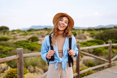 Sırt çantalı, şapkalı, ahşap patikada yürüyen genç bir kadın gezginin arka görüntüsü. Doğanın tadını çıkaran güzel bir kadın, deniz manzarası. Yürüyüş, yürüyüş konsepti.