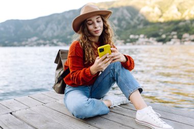Güneş doğarken elinde telefonla deniz manzarasının tadını çıkaran şapkalı genç bir kadın. Şirin gezgin blogları, mesajlaşmaları, fotoğraf çekmeleri. Seyahat, özgürlük ve yürüyüş konsepti. Etkin yaşam biçimi.