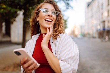 Şık giyinmiş, gözlüklü, elinde telefonla güneşli caddelerde yürüyen mutlu turist portresi. Dışarıda blog yazan güzel bir kadın. Bloglama, yürüme konsepti.