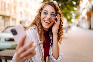 Şık giyinmiş, gözlüklü, elinde telefonla güneşli caddelerde yürüyen mutlu turist portresi. Dışarıda blog yazan güzel bir kadın. Bloglama, yürüme konsepti.