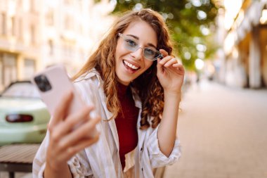 Şık giyinmiş, gözlüklü, elinde telefonla güneşli caddelerde yürüyen mutlu turist portresi. Dışarıda blog yazan güzel bir kadın. Bloglama, yürüme konsepti.