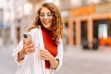 Şık giyinmiş, gözlüklü, elinde telefonla güneşli caddelerde yürüyen mutlu turist portresi. Dışarıda blog yazan güzel bir kadın. Bloglama, yürüme konsepti.