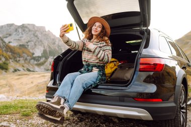 Telefonu olan genç bir kadın bir arabanın bagajında otururken dağ manzarasının tadını çıkarıyor. Güzel bir gezgin araba macerası hakkında blog yazıyor. Seyahat, özgürlük konsepti. Etkin yaşam biçimi.