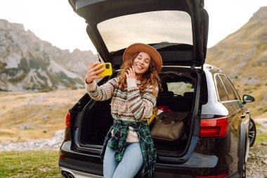 Telefonu olan genç bir kadın bir arabanın bagajında otururken dağ manzarasının tadını çıkarıyor. Güzel bir gezgin araba macerası hakkında blog yazıyor. Seyahat, özgürlük konsepti. Etkin yaşam biçimi.