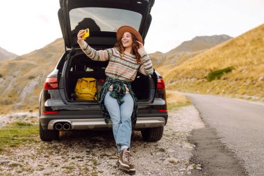 Telefonu olan genç bir kadın bir arabanın bagajında otururken dağ manzarasının tadını çıkarıyor. Güzel bir gezgin araba macerası hakkında blog yazıyor. Seyahat, özgürlük konsepti. Etkin yaşam biçimi.