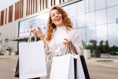 Alışveriş merkezinin yakınında alışveriş torbaları taşıyan genç bir kadın. Neşeli kadın dışarıda alışveriş yaptıktan sonra eğleniyor. Alışveriş konsepti, tüketim. Etkin yaşam biçimi.