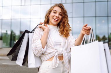 Alışveriş merkezinin yakınında alışveriş torbaları taşıyan genç bir kadın. Neşeli kadın dışarıda alışveriş yaptıktan sonra eğleniyor. Alışveriş konsepti, tüketim. Etkin yaşam biçimi.