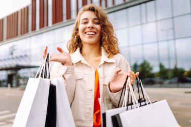 Alışveriş merkezinin yakınında alışveriş torbaları taşıyan genç bir kadın. Neşeli kadın dışarıda alışveriş yaptıktan sonra eğleniyor. Alışveriş konsepti, tüketim. Etkin yaşam biçimi.