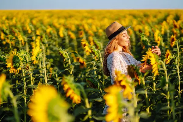Elbise giymiş güzel bir kadın ayçiçeği tarlasında sıcak bir günbatımının tadını çıkarıyor. Genç bir kadın açık havada yürür ve özgürlüğü hisseder. Yürüme konsepti. Etkin yaşam biçimi.