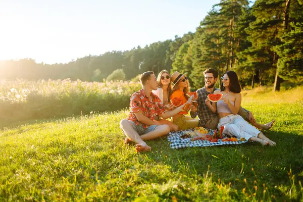 Mutlu arkadaşlar piknikte karpuz yiyorlar. Genç kadınlar ve erkekler gün batımında birlikte dışarıda vakit geçiriyorlar. Arkadaşlık kavramı, eğlence..