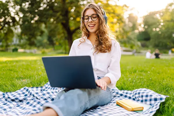 Yeşil çimenlikte çalışan, kablosuz kulaklıklı ve dizüstü bilgisayarlı sıradan giyinmiş mutlu bir kadın. Genç bir kız öğrenci ya da serbest çalışan parkta dizüstü bilgisayarla havanın tadını çıkarıyor.. 