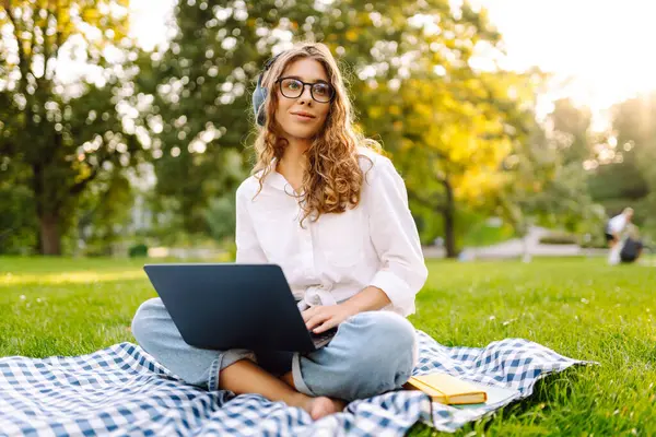 Yeşil çimenlikte çalışan, kablosuz kulaklıklı ve dizüstü bilgisayarlı sıradan giyinmiş mutlu bir kadın. Genç bir kız öğrenci ya da serbest çalışan parkta dizüstü bilgisayarla havanın tadını çıkarıyor.. 