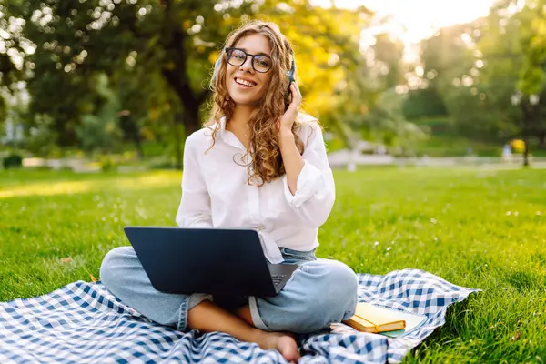 Yeşil çimenlikte çalışan, kablosuz kulaklıklı ve dizüstü bilgisayarlı sıradan giyinmiş mutlu bir kadın. Genç bir kız öğrenci ya da serbest çalışan parkta dizüstü bilgisayarla havanın tadını çıkarıyor.. 