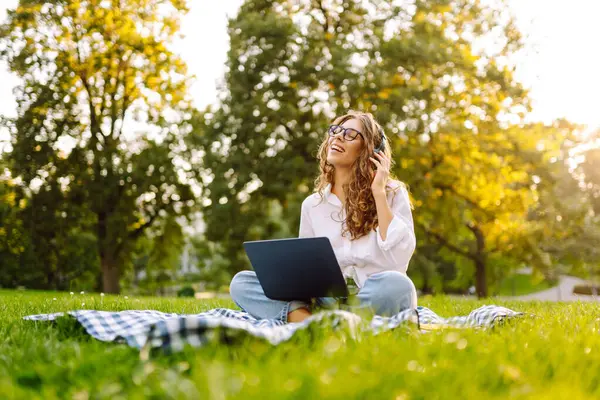 Yeşil çimenlikte çalışan, kablosuz kulaklıklı ve dizüstü bilgisayarlı sıradan giyinmiş mutlu bir kadın. Genç bir kız öğrenci ya da serbest çalışan parkta dizüstü bilgisayarla havanın tadını çıkarıyor.. 