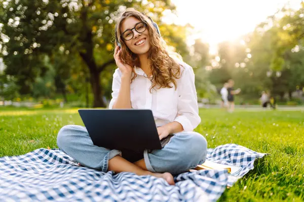 Yeşil çimenlikte çalışan, kablosuz kulaklıklı ve dizüstü bilgisayarlı sıradan giyinmiş mutlu bir kadın. Genç bir kız öğrenci ya da serbest çalışan parkta dizüstü bilgisayarla havanın tadını çıkarıyor.. 