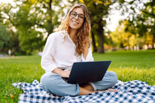 Yeşil çimenlikte çalışan, kablosuz kulaklıklı ve dizüstü bilgisayarlı sıradan giyinmiş mutlu bir kadın. Genç bir kız öğrenci ya da serbest çalışan parkta dizüstü bilgisayarla havanın tadını çıkarıyor.. 