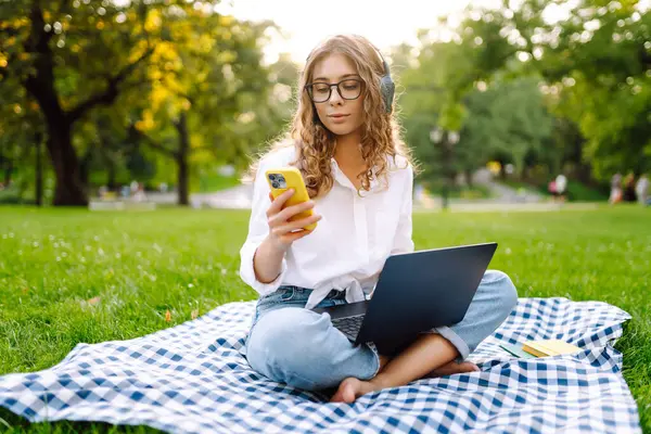 Yeşil çimenlikte çalışan, kablosuz kulaklıklı ve dizüstü bilgisayarlı sıradan giyinmiş mutlu bir kadın. Genç bir kız öğrenci ya da serbest çalışan parkta dizüstü bilgisayarla havanın tadını çıkarıyor.. 