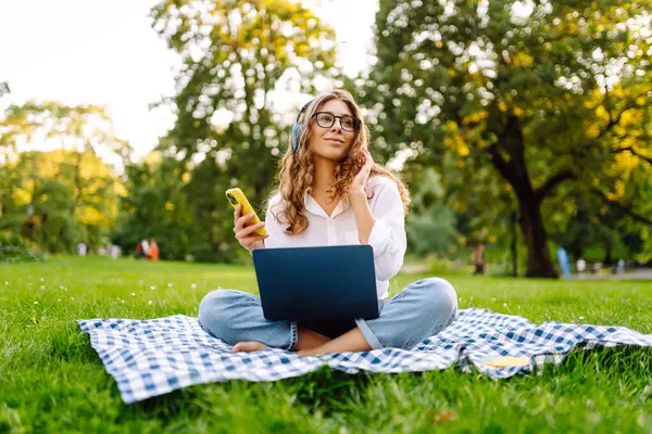 Yeşil çimenlikte çalışan, kablosuz kulaklıklı ve dizüstü bilgisayarlı sıradan giyinmiş mutlu bir kadın. Genç bir kız öğrenci ya da serbest çalışan parkta dizüstü bilgisayarla havanın tadını çıkarıyor.. 