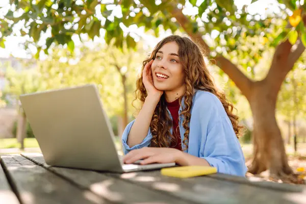 Parktaki bir masada dizüstü bilgisayarla çalışan genç bir kadın. Açık havada havanın tadını çıkaran mutlu kadın serbest yazar. Uzaktan çalışma konsepti. Yaşam biçimi.