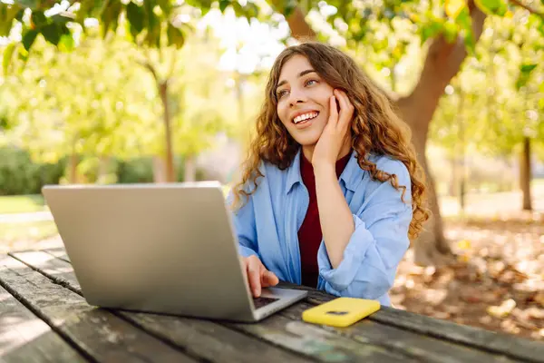 Parktaki bir masada dizüstü bilgisayarla çalışan genç bir kadın. Açık havada havanın tadını çıkaran mutlu kadın serbest yazar. Uzaktan çalışma konsepti. Yaşam biçimi.