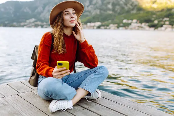 Güneş doğarken elinde telefonla deniz manzarasının tadını çıkaran şapkalı genç bir kadın. Şirin gezgin blogları, mesajlaşmaları, fotoğraf çekmeleri. Seyahat, özgürlük ve yürüyüş konsepti. Etkin yaşam biçimi.