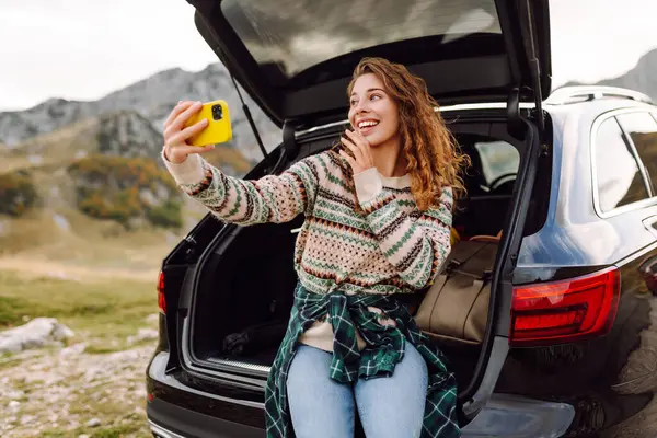 Telefonu olan genç bir kadın bir arabanın bagajında otururken dağ manzarasının tadını çıkarıyor. Güzel bir gezgin araba macerası hakkında blog yazıyor. Seyahat, özgürlük konsepti. Etkin yaşam biçimi.