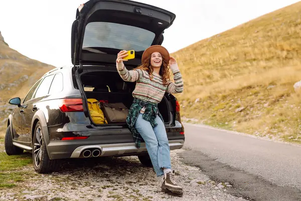 Telefonu olan genç bir kadın bir arabanın bagajında otururken dağ manzarasının tadını çıkarıyor. Güzel bir gezgin araba macerası hakkında blog yazıyor. Seyahat, özgürlük konsepti. Etkin yaşam biçimi.