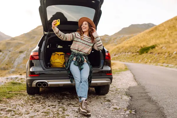 Telefonu olan genç bir kadın bir arabanın bagajında otururken dağ manzarasının tadını çıkarıyor. Güzel bir gezgin araba macerası hakkında blog yazıyor. Seyahat, özgürlük konsepti. Etkin yaşam biçimi.