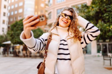 Telefonlu, güneş gözlüklü güzel bir turist gün batımında selfie çekiyor. Genç bir kadın dışarıda yürümekten hoşlanıyor. Blogculuk, turizm, eğlence kavramı.