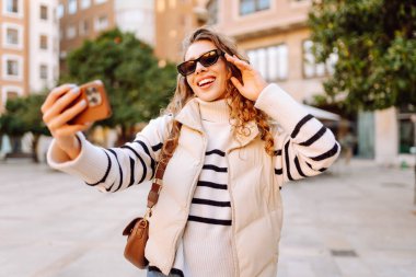 Telefonlu, güneş gözlüklü güzel bir turist gün batımında selfie çekiyor. Genç bir kadın dışarıda yürümekten hoşlanıyor. Blogculuk, turizm, eğlence kavramı.