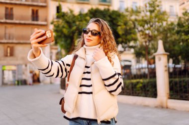 Telefonlu, güneş gözlüklü güzel bir turist gün batımında selfie çekiyor. Genç bir kadın dışarıda yürümekten hoşlanıyor. Blogculuk, turizm, eğlence kavramı.