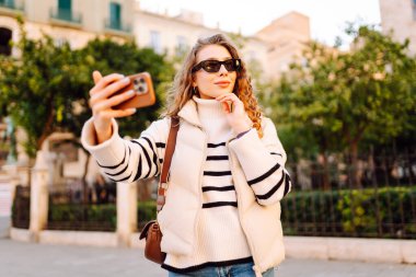 Telefonlu, güneş gözlüklü güzel bir turist gün batımında selfie çekiyor. Genç bir kadın dışarıda yürümekten hoşlanıyor. Blogculuk, turizm, eğlence kavramı.